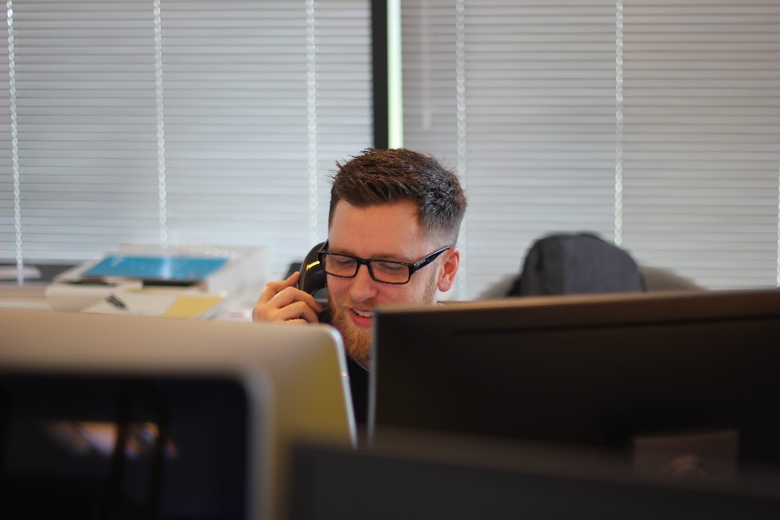 A man holds a home phone up to his ear while making a long-distance call. He is sitting behind two monitor screens and looks happy to hear the voice on the other end of the line.